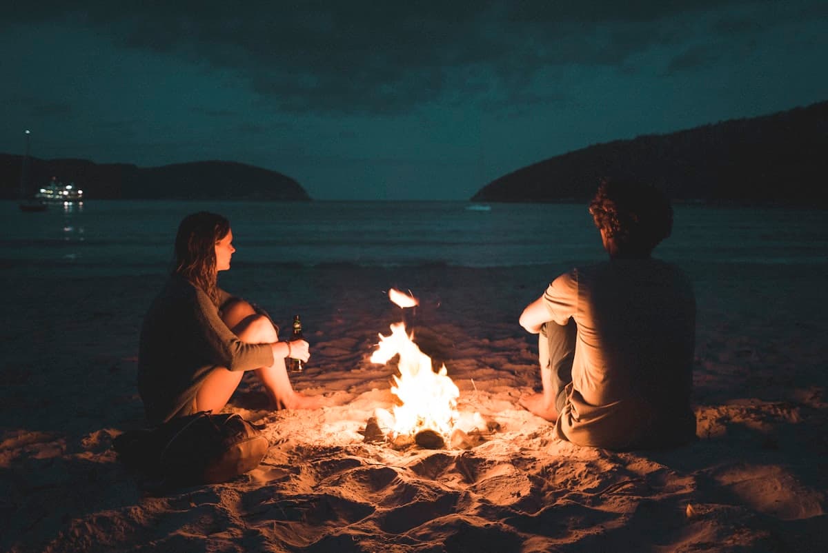 Two people sitting close by a campfire on a beach at night