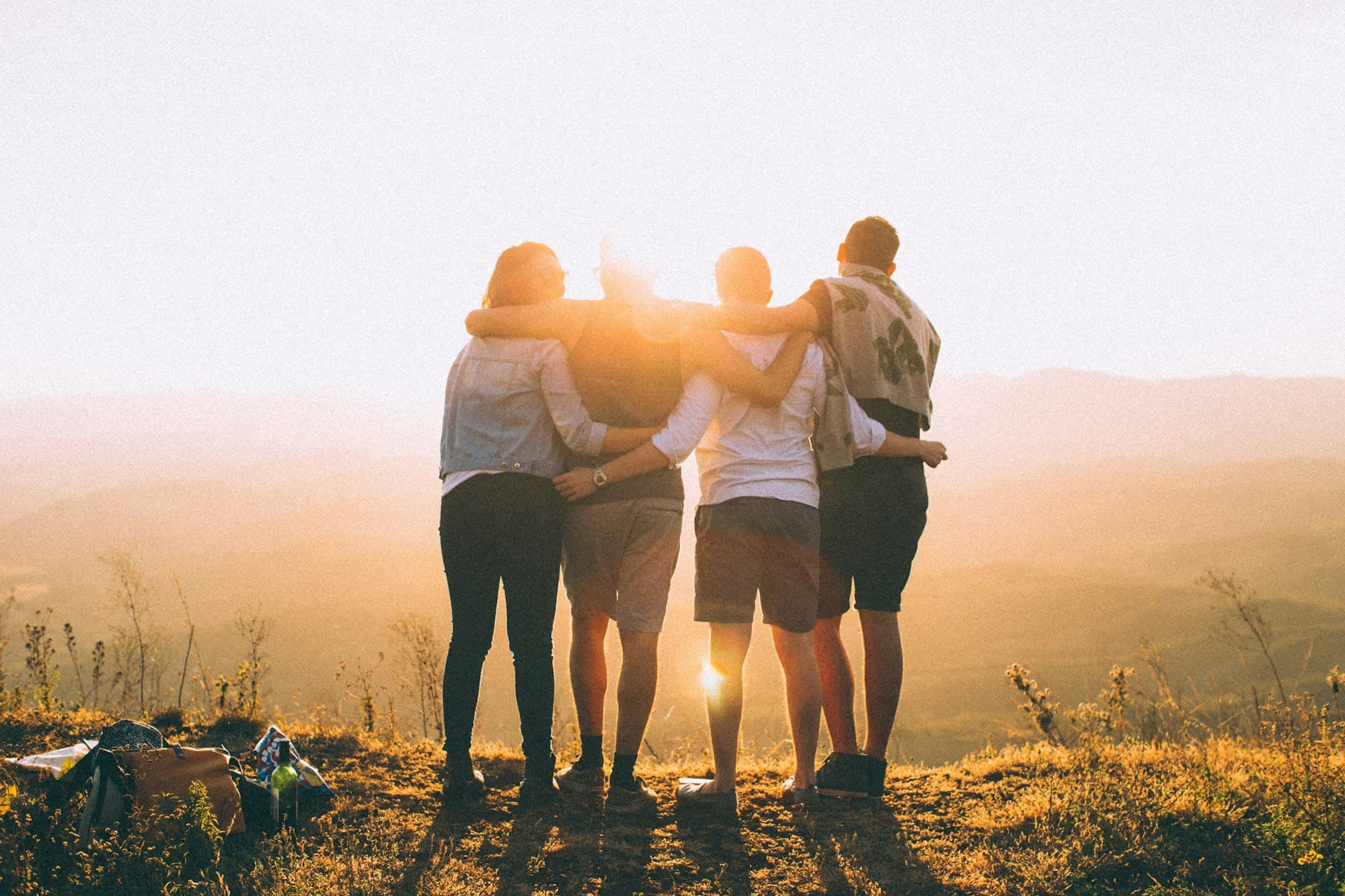 Four friends standing arm in arm on a hilltop, facing the sunset