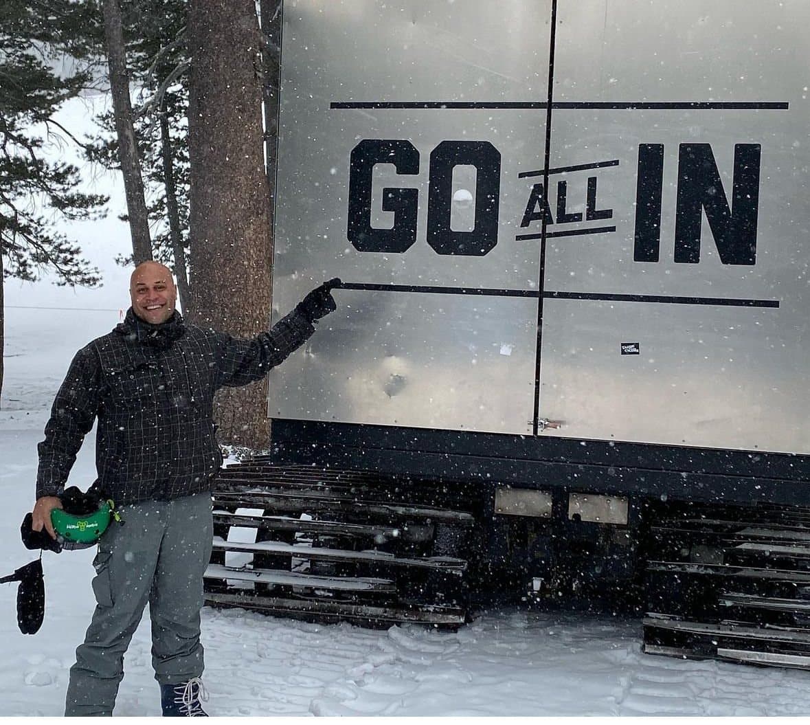 Jonathan in snowboarding gear pointing at a trailer painted GO ALL IN